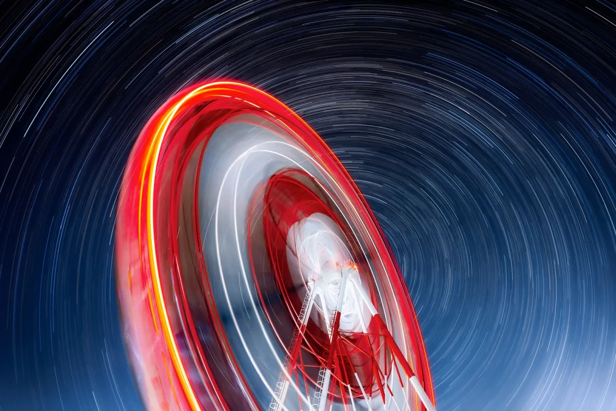 Image showing long exposure from ground level looking up at a red and white ferris wheel with circular star trails behind